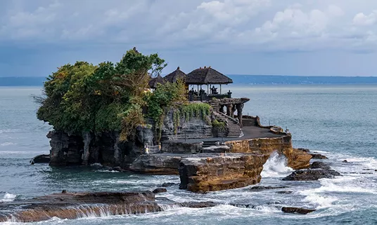 Ancient Balinese sea temple perched on a rocky coastal outcrop surrounded by crashing ocean waves under a dramatic cloudy sky.
