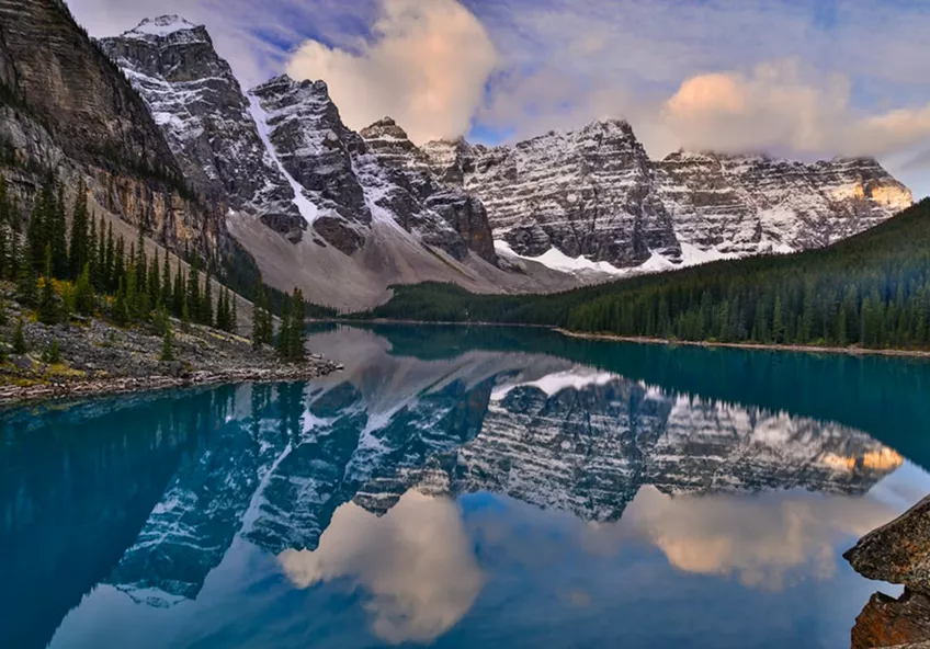 Snow-capped mountains perfectly reflected in the turquoise waters of Moraine Lake, surrounded by pine forests.