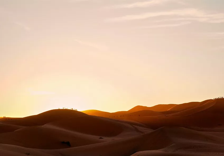 Silhouettes of people standing on rolling sand dunes against a hazy, glowing orange sunset sky.