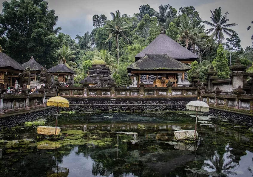 An ancient Balinese water temple surrounded by lush tropical trees and a reflective green pond.