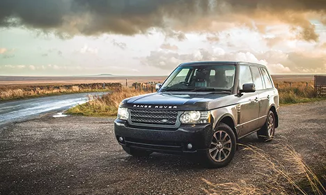 Dark blue Range Rover SUV parked on a gravel road with dramatic cloudy skies and open moorland behind it.