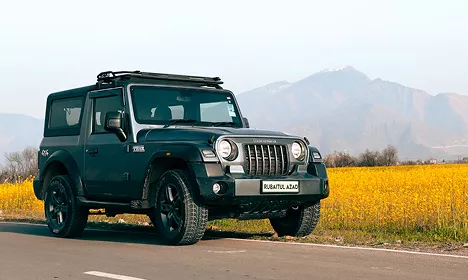 Dark green Mahindra Thar 4x4 parked on a road beside yellow mustard fields with mountains in the background.