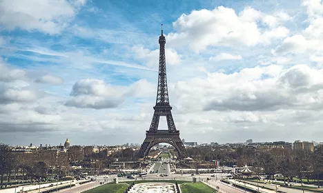 The Eiffel Tower standing tall against a partly cloudy sky, viewed from the Champ de Mars gardens in Paris.