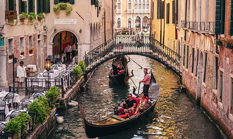 Gondolas carrying tourists through a narrow Venice canal lined with colourful buildings and a stone bridge overhead.