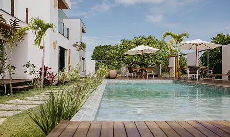 Outdoor pool with a wooden deck, white umbrellas, and tropical plants beside a modern white villa under a blue sky.