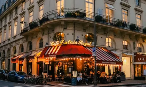 Café Palais Royal in Paris at dusk, with red awnings, glowing lights, and guests dining at outdoor pavement tables.