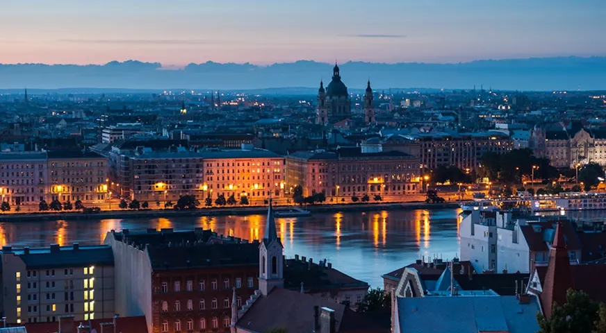 A wide cityscape at dusk showing historic European architecture and a river reflecting the golden lights of the buildings.