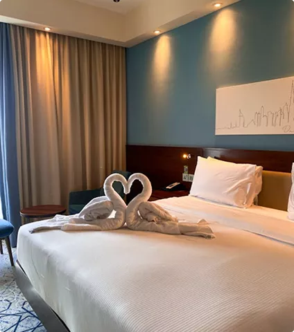 A luxury hotel room featuring a bed with two towels folded into a heart-shaped swan design and soft blue accent lighting.