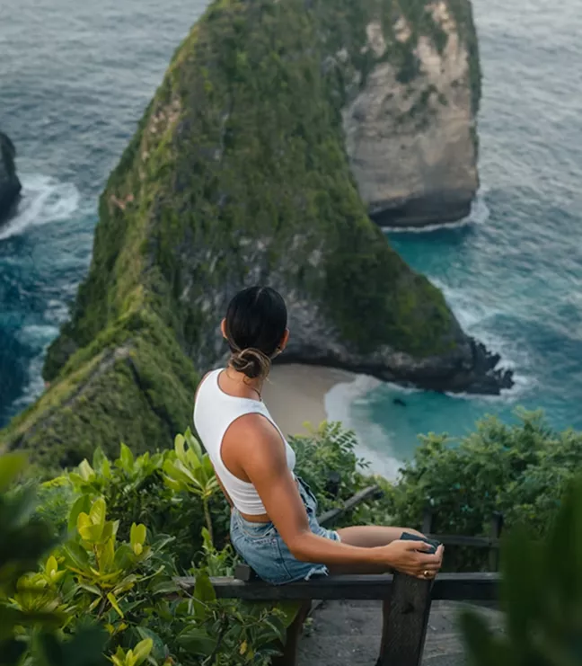 A woman sits on a wooden railing overlooking the iconic T-Rex shaped Kelingking Beach cliff and turquoise water in Nusa Penida, Bali.