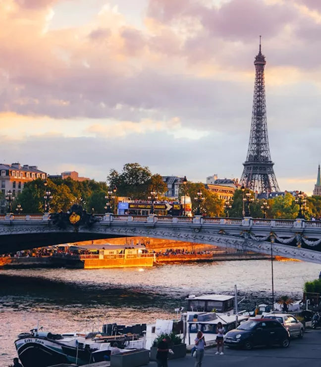 The Eiffel Tower rises above the Seine River in Paris at sunset, with a stone bridge and tour boats visible in the glowing evening light.