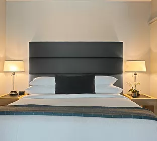 Warmly lit hotel room with a king bed, charcoal padded headboard, white pillows, and matching bedside lamps with orchids.