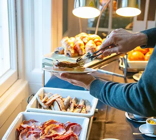 Guest serving food at a hotel breakfast buffet with pastries, bacon, and warm dishes under heat lamps near a bright window.