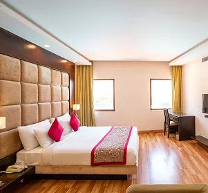 Hotel room with a king bed, quilted gold headboard, hot-pink accent pillows, hardwood floors, and a work desk near the window.