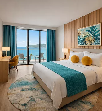 Bright hotel room with a king bed, teal accents, tropical artwork, and a balcony overlooking a blue ocean bay.