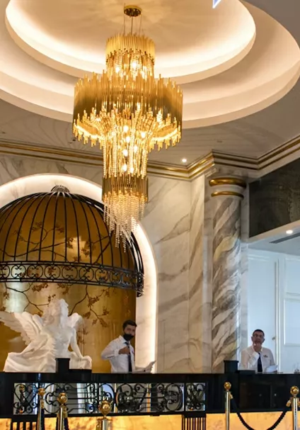 An ornate hotel reception area with a massive gold tiered chandelier, marble columns, and a decorative gold birdcage structure behind the desk.