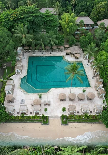 Aerial view of a large resort swimming pool surrounded by sun loungers, umbrellas, lush tropical greenery, and a sandy beachfront.