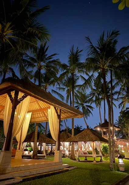 Illuminated Balinese thatched gazebos with white curtains glowing warmly at night surrounded by tall tropical palm trees under a starry sky.