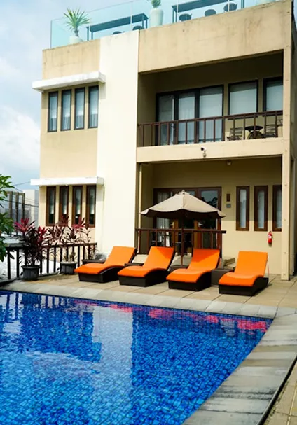 A hotel outdoor pool with bright orange sun loungers and a beige umbrella in front of a modern multi-storey building facade.