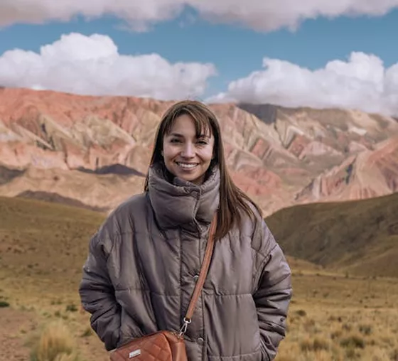 A smiling woman in a brown puffer jacket and crossbody bag stands in a vast, mountainous desert landscape under a blue sky.