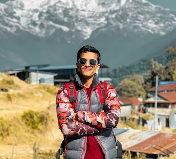 A man in sunglasses and a camouflage puffer vest stands with arms crossed against a backdrop of snow-capped mountains.