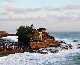 Tanah Lot sea temple standing on a rocky island at sunset, with waves crashing around it and visitors gathered on the shoreline.