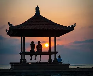 Silhouette of two people sitting in a traditional Balinese pavilion gazebo watching a vibrant golden sunset over the calm ocean.