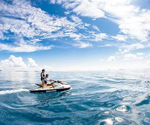 Person riding a jet ski across calm blue ocean under a bright sky with scattered clouds, leaving a trail of ripples behind.