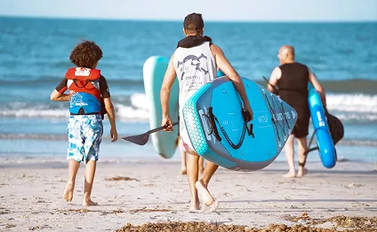 A family walks across a sandy beach toward the ocean, carrying bright blue paddleboards and oars for a day of water activities.