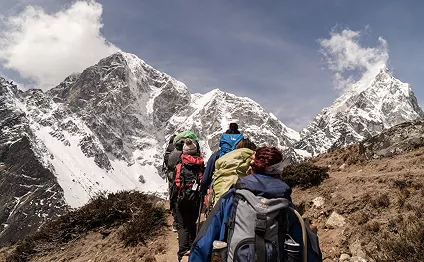 A line of hikers with heavy backpacks follows a dirt trail toward towering, snow-covered Himalayan peaks under a clear blue sky.