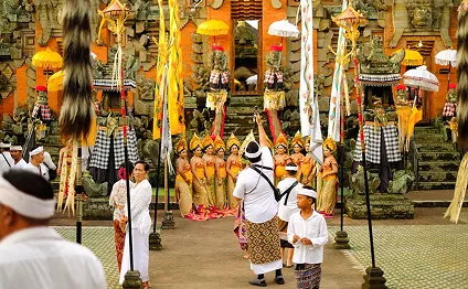 Balinese performers in traditional gold costumes line up during a colorful Hindu temple ceremony surrounded by ornate stone carvings.