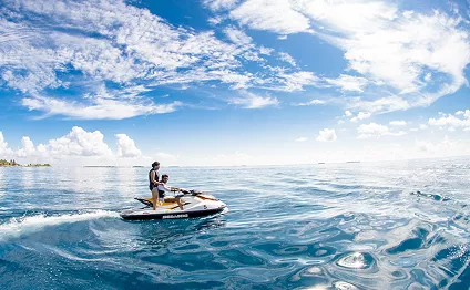 A person riding a jet ski across a calm, crystal-blue tropical ocean under a wide blue sky with scattered white clouds.