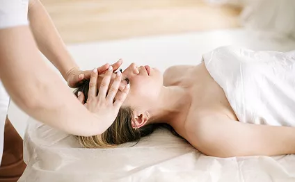 A therapist gently massages a relaxed woman's head and face as she lies on a spa treatment table draped in soft white linen.